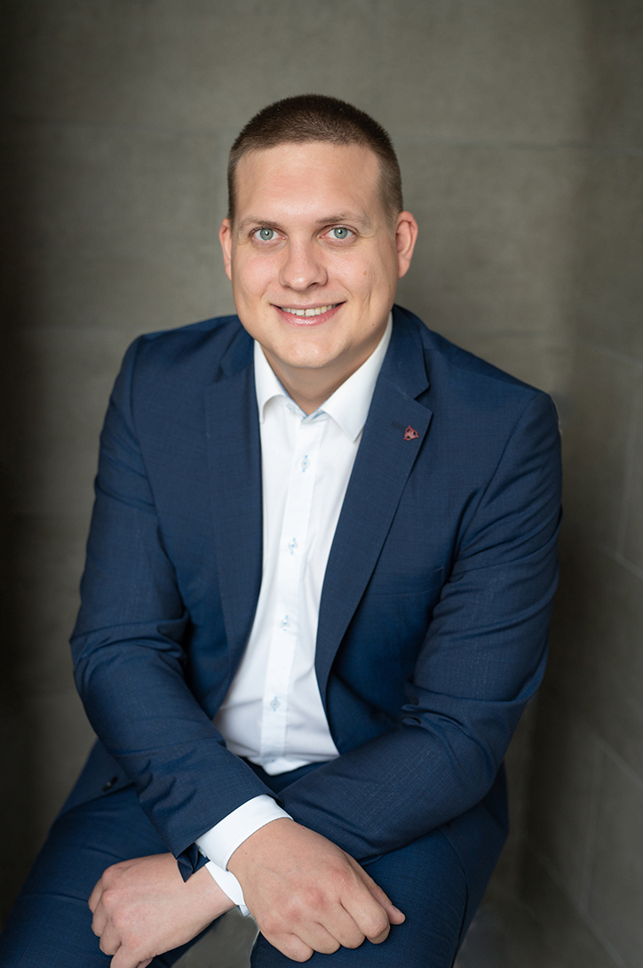 Porträtfoto Marius Gehrmann, junger Mann in dunkelblauem Anzug und weißem Hemd vor einer grauen Wand freundlich lächelnd.